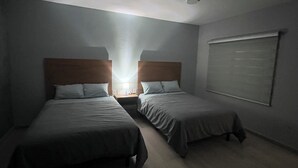 A hotel room with two single beds, a nightstand with a lamp, a window with blinds, and a ceiling light.