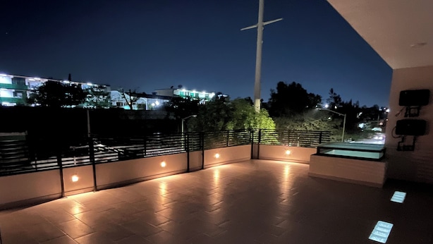 A rooftop terrace with a railing, a cross, and a view of buildings in the background.