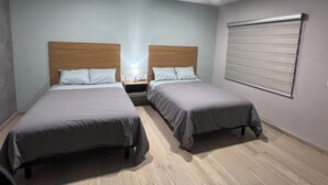 A hotel room with two single beds, a nightstand with a lamp, a window with blinds, and a ceiling light.