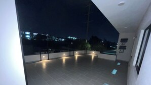 A spacious balcony with tiled flooring, a railing, and a view of city lights in the background.