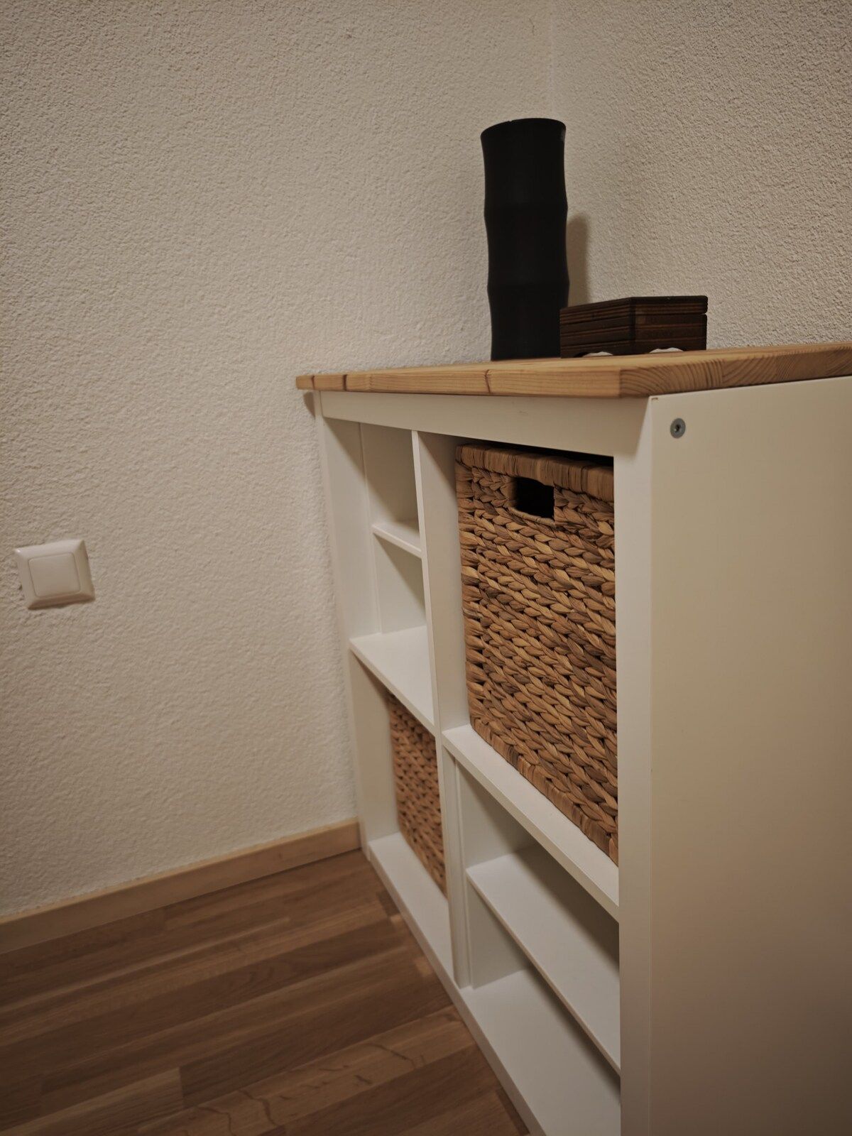 Un coin de pièce avec un mur blanc, un plancher en bois et une étagère blanche sur laquelle se trouve un panier en osier.