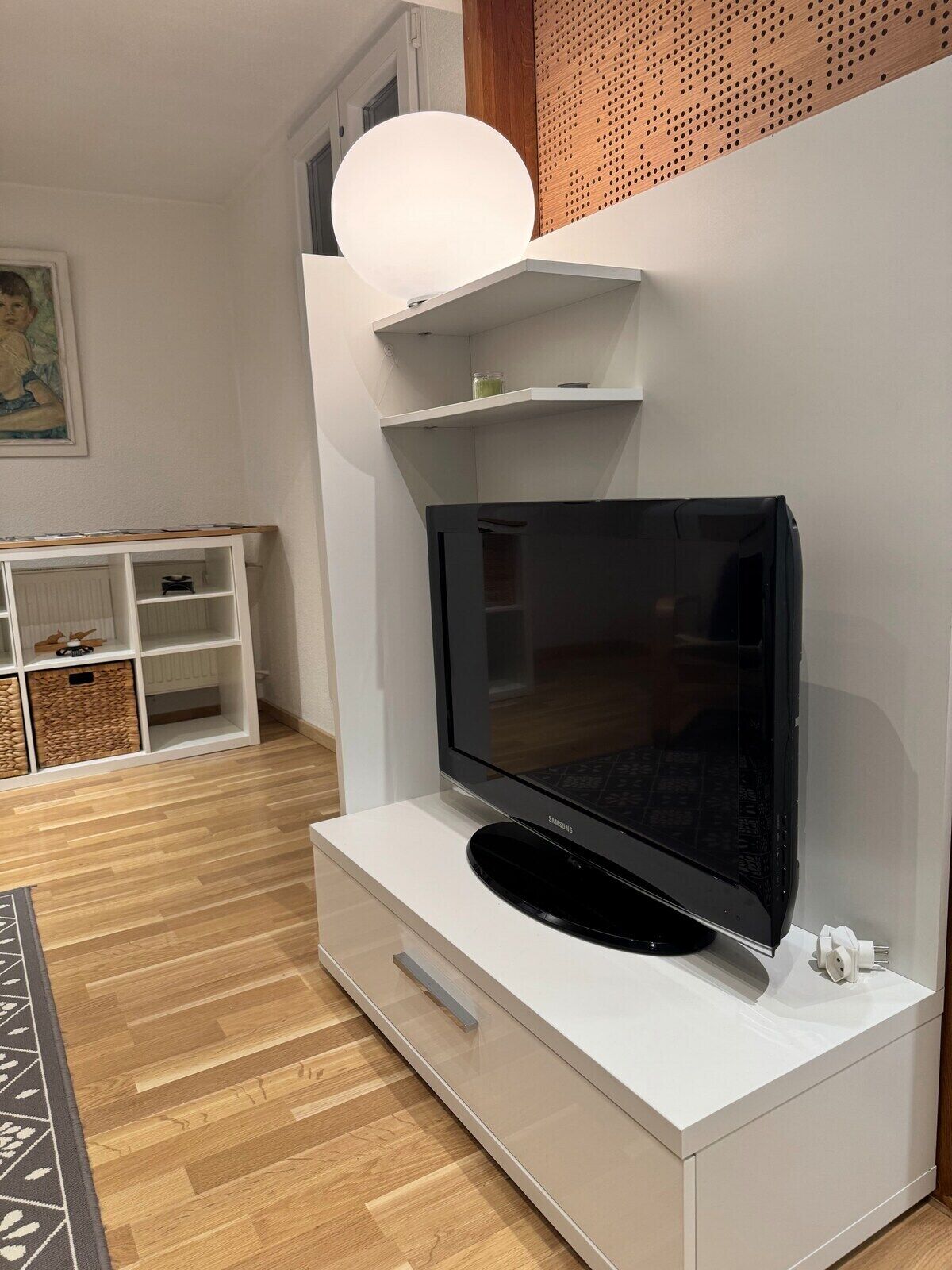 Una sala de estar moderna con un televisor de pantalla plana montado en un mueble blanco, piso de madera y una pared blanca con un estante decorativo.
