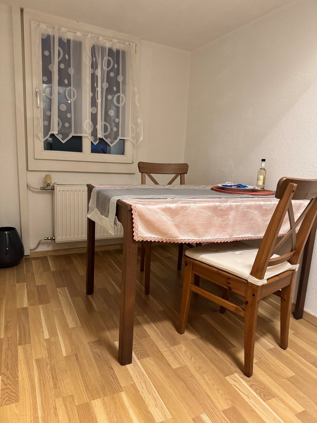 Ruokailutila, jossa on kahdelle katettu pöytä, ikkuna verhoilla, patteri ja puulattia.