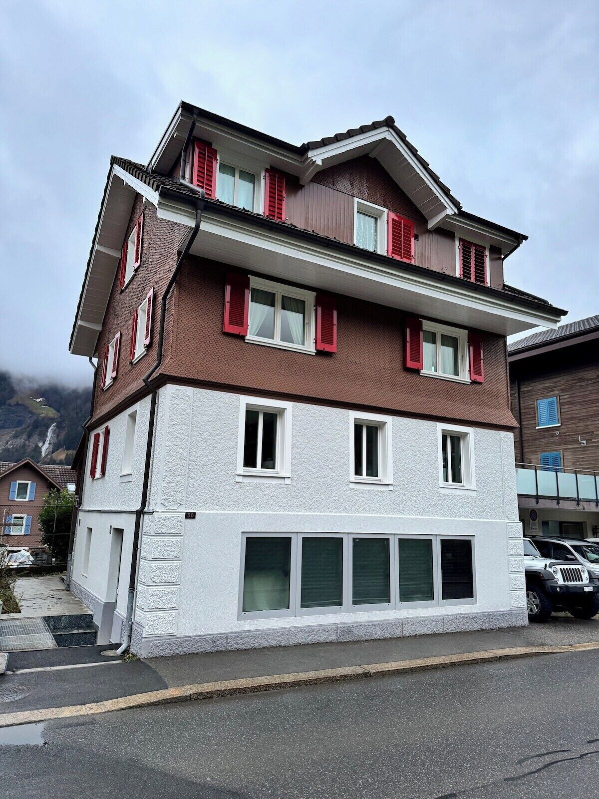 Un immeuble à plusieurs étages, doté d’une façade blanche et de volets rouges, avec une entrée de garage et un espace de stationnement.