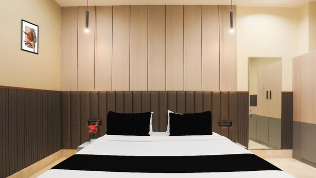 A hotel room with a large bed, a wooden headboard, a bedside table with a vase, and a mirror. - Deluxe Hotel Heritage Plaza (Hisar)