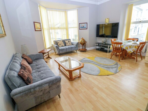 A living room with a sofa, a coffee table, a TV, a dining table with chairs, and a window with curtains. - 1 Hilton Flats (Tenby)