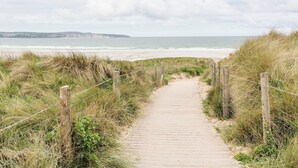 Ein Holzweg, der zu einem Strand mit grasbewachsenen Dünen und einem Zaun an einer Seite führt.
