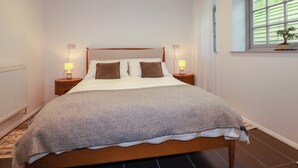 Ein ordentlich bezogenes Bett mit grauer Decke, zwei Nachttischlampen, ein Fenster mit Jalousien und eine Heizung.