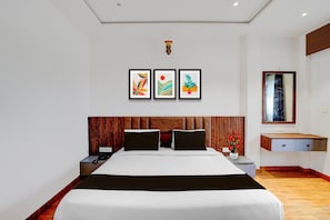 A modern hotel room with a large bed, a wooden headboard, a bedside table with a lamp, a small desk, and a mirror.
