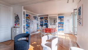 Een ruime woonkamer met een grote boekenkast, een eethoek en uitzicht op het groen.