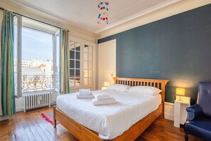Ein Schlafzimmer mit einem hölzernen Bett, Blick auf die Stadt durch ein Fenster und ein Balkon mit Geländer.