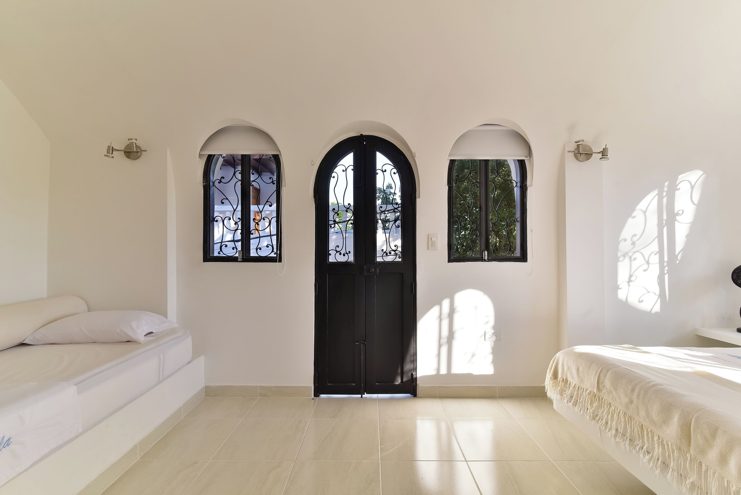 Una habitación con una cama, una puerta y dos ventanas con detalles de hierro forjado.