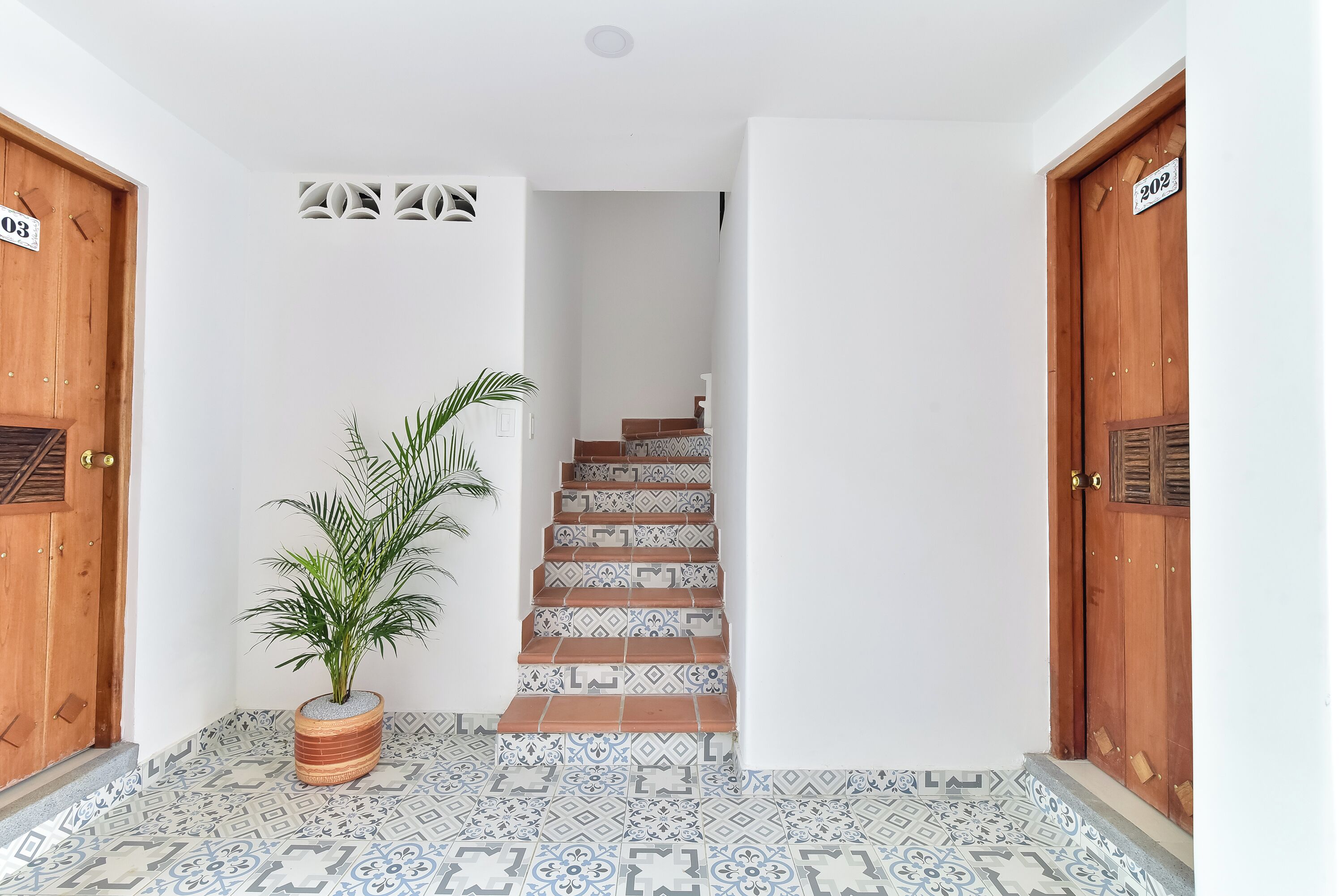A hallway with a wooden door labeled '03', a potted plant, patterned floor tiles, and a staircase with patterned tiles leading to another room.