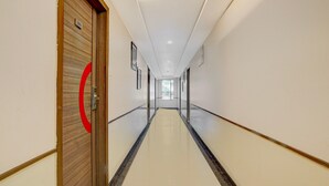 A long, narrow hallway with a wooden door on the left, a red circular sign, and a window at the end. - Deluxe Hotel Kapil Residency (Pune)
