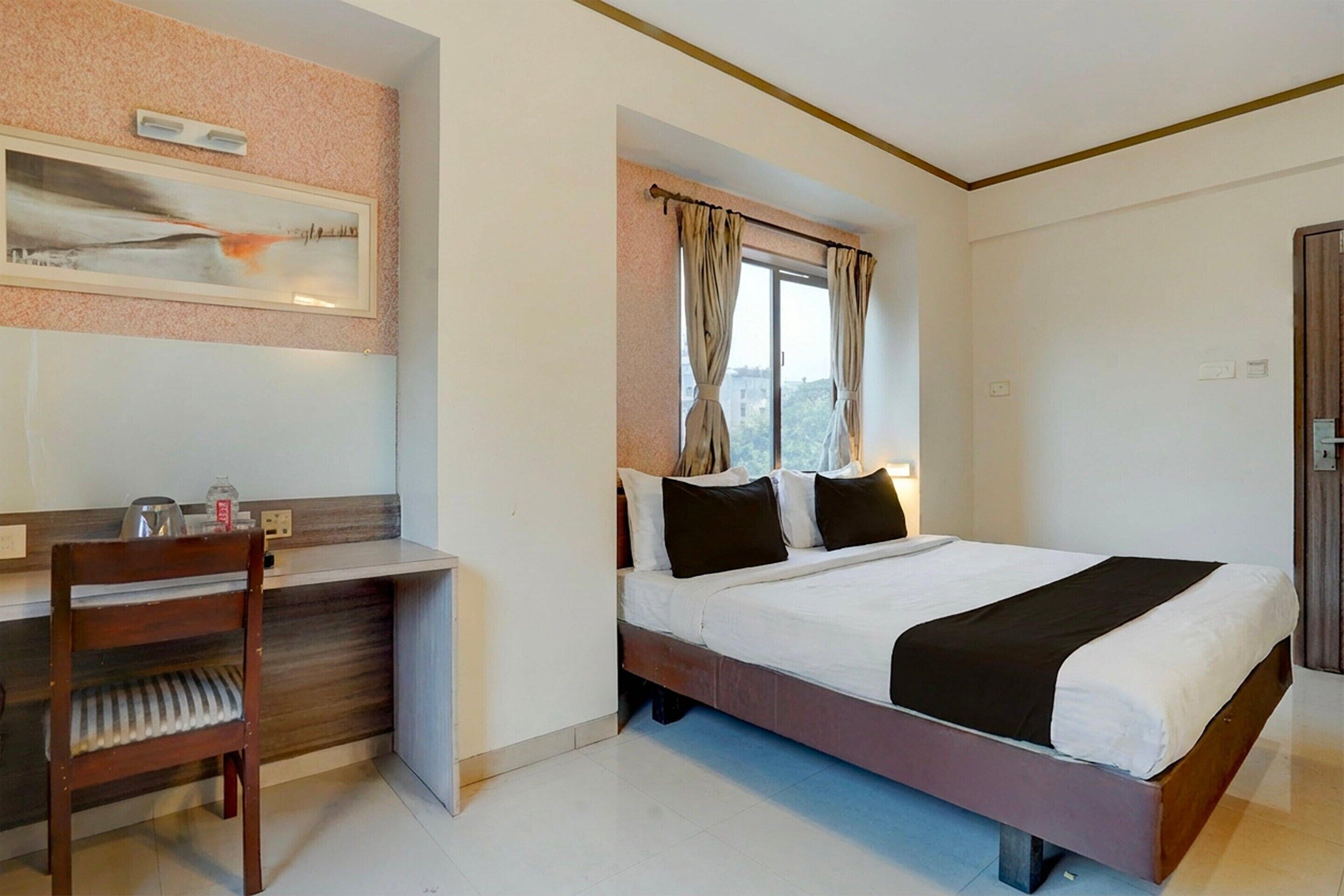 Une chambre d’hôtel comprenant un lit, un bureau, une chaise, une fenêtre avec des rideaux et une étagère fixée au mur, sur laquelle est posée une image.
