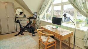A bedroom with a desk, chair, exercise bike, and a large window with curtains. - Lakes View (Keswick)