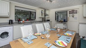 A modern kitchen with a dining table set for six, a washing machine, and a large window with a view. - Preswylfa (Cemaes Bay)