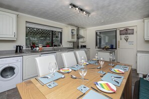 A modern kitchen with a dining table set for six, a washing machine, and a large window with a view. - Preswylfa (Cemaes Bay)