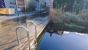 Un patio in legno con ringhiere metalliche che conduce a una superficie d'acqua tranquilla, una cucina moderna e un'area pranzo con divano e sedie.