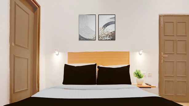 A bed with a wooden headboard, two black pillows, a white bedspread, and a small bedside table with a plant. - Deluxehotel Woodland (Nagpur)