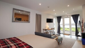 A bedroom with a bed, a mirror, a television, a table with chairs, and a view of the outdoors. - Faodail Studio (Skeabost)