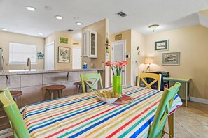 A dining area with a striped tablecloth, chairs, a vase with flowers, and a kitchen area with a window and a cabinet. - Summerhouse Townhomes #2- Changes in Attitude (Miramar Beach)