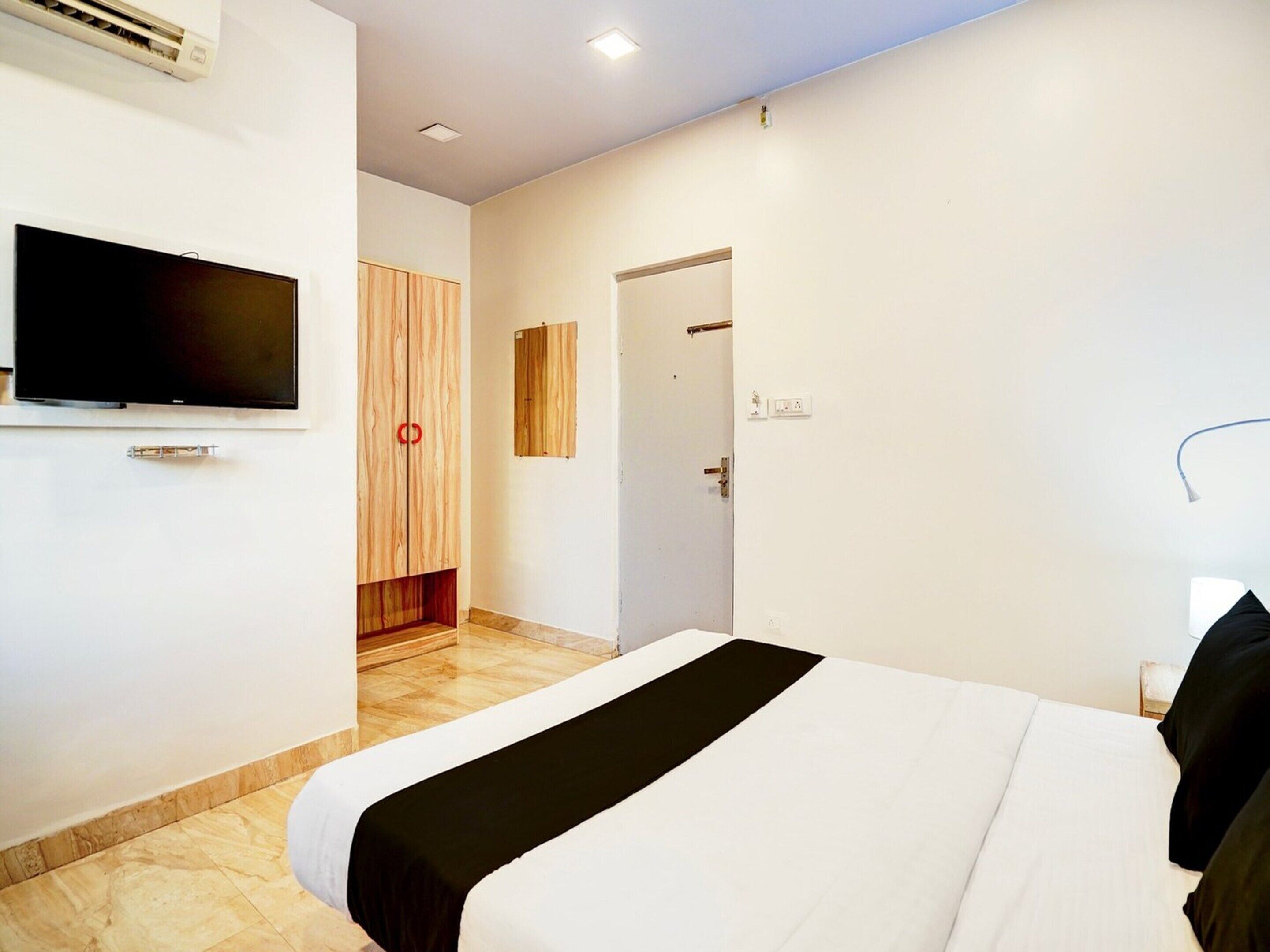 A hotel room with a bed, a television mounted on the wall, a wooden wardrobe, and a door leading to another room.