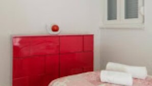 Une chambre avec une tête de lit rouge, une fenêtre avec des stores et un lit recouvert de draps blancs.