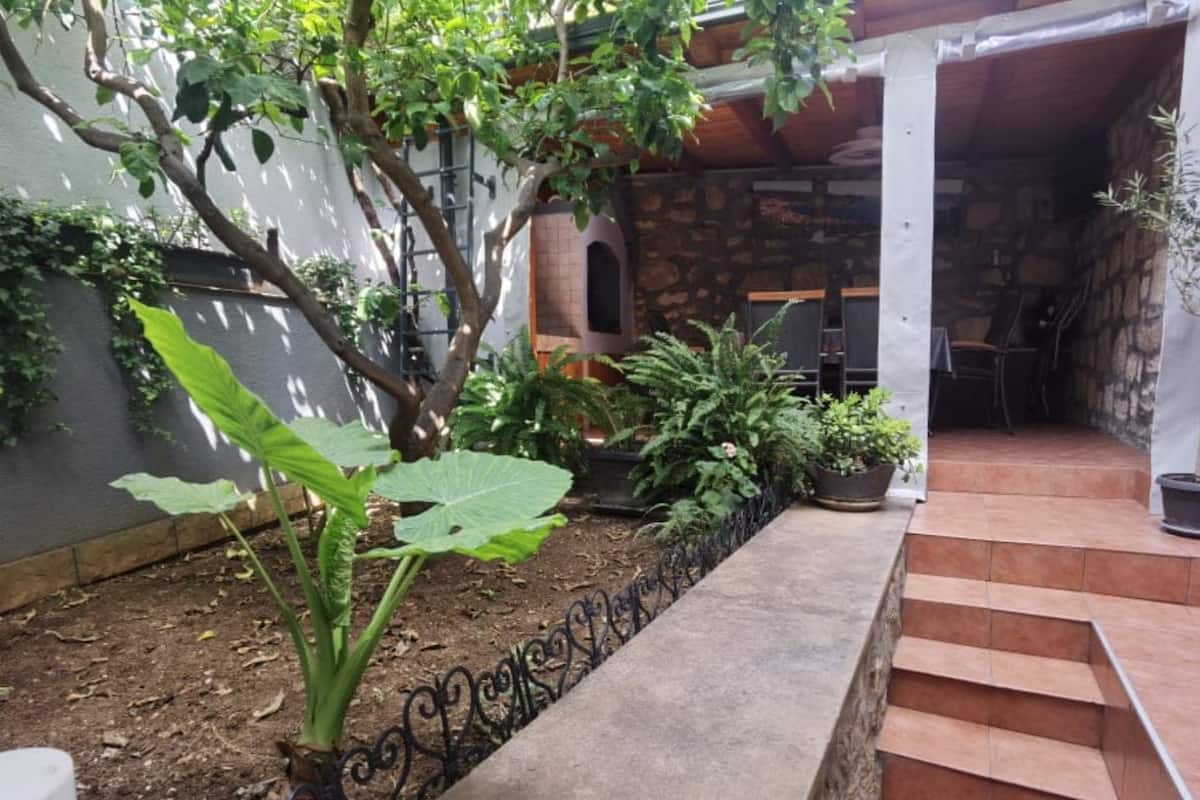 A stone building with a staircase leading to a covered area, surrounded by greenery and a metal fence.