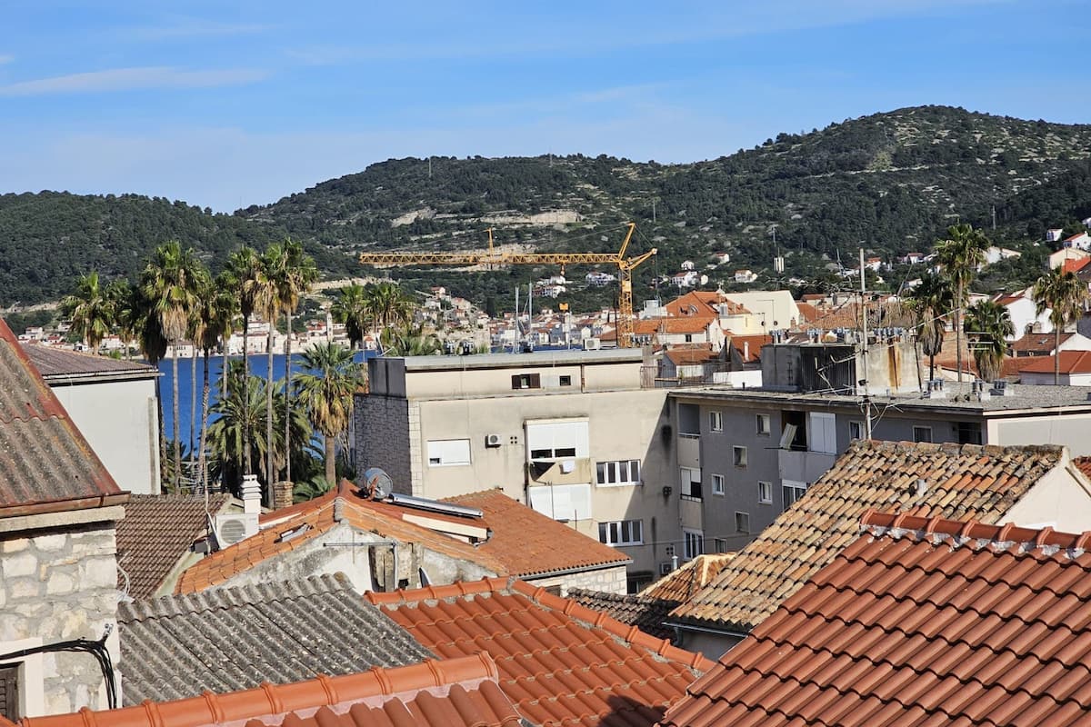 A coastal cityscape with terracotta-roofed buildings, palm trees, and a distant construction crane.
