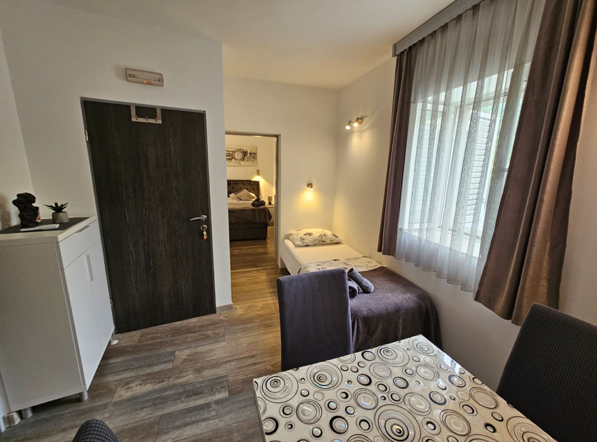 Et lille hotelværelse med seng, skrivebord, stol, bord og et vindue med gardiner.