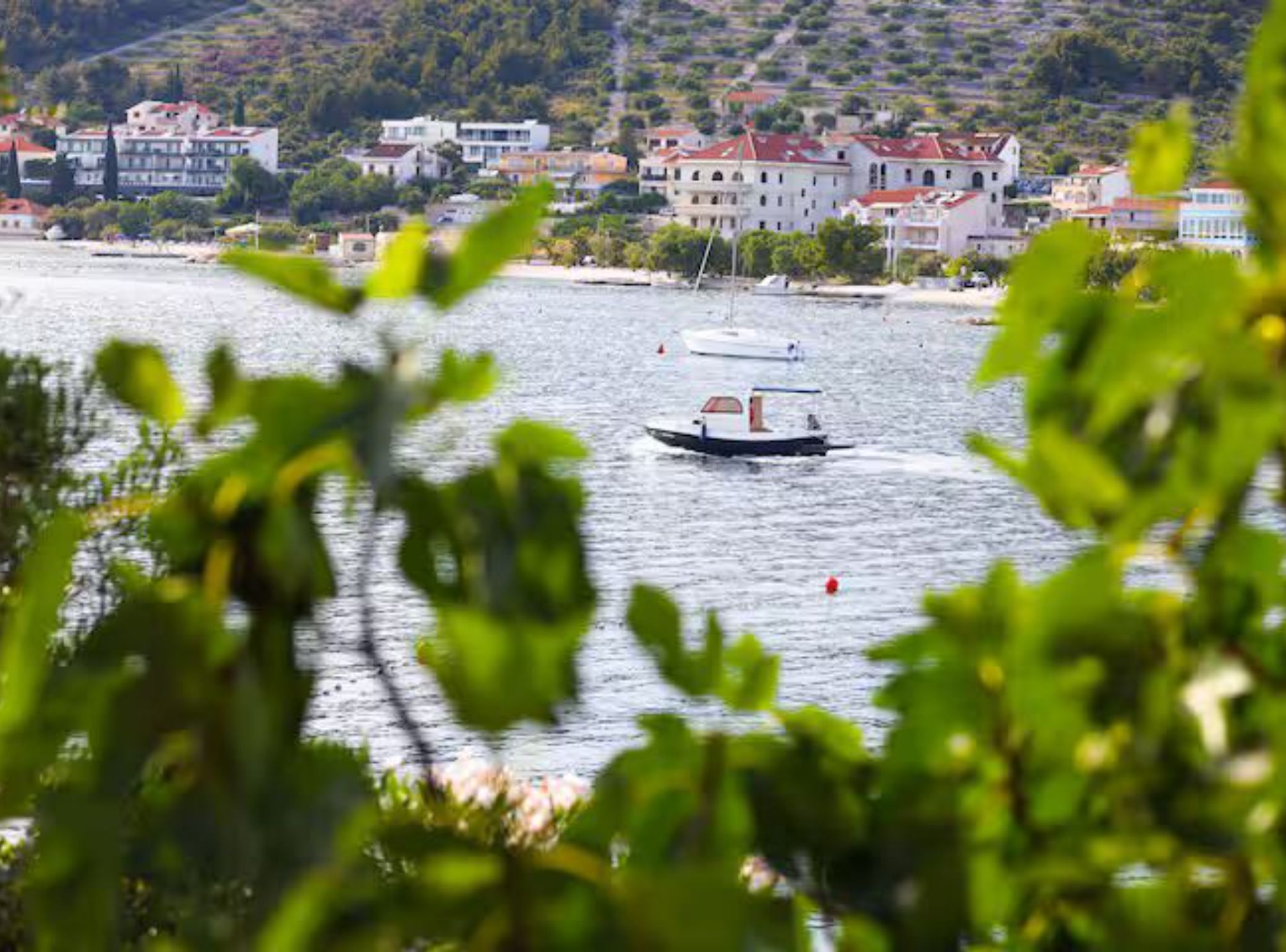 A coastal town with buildings along the waterfront, a boat on the water, and greenery in the foreground.
