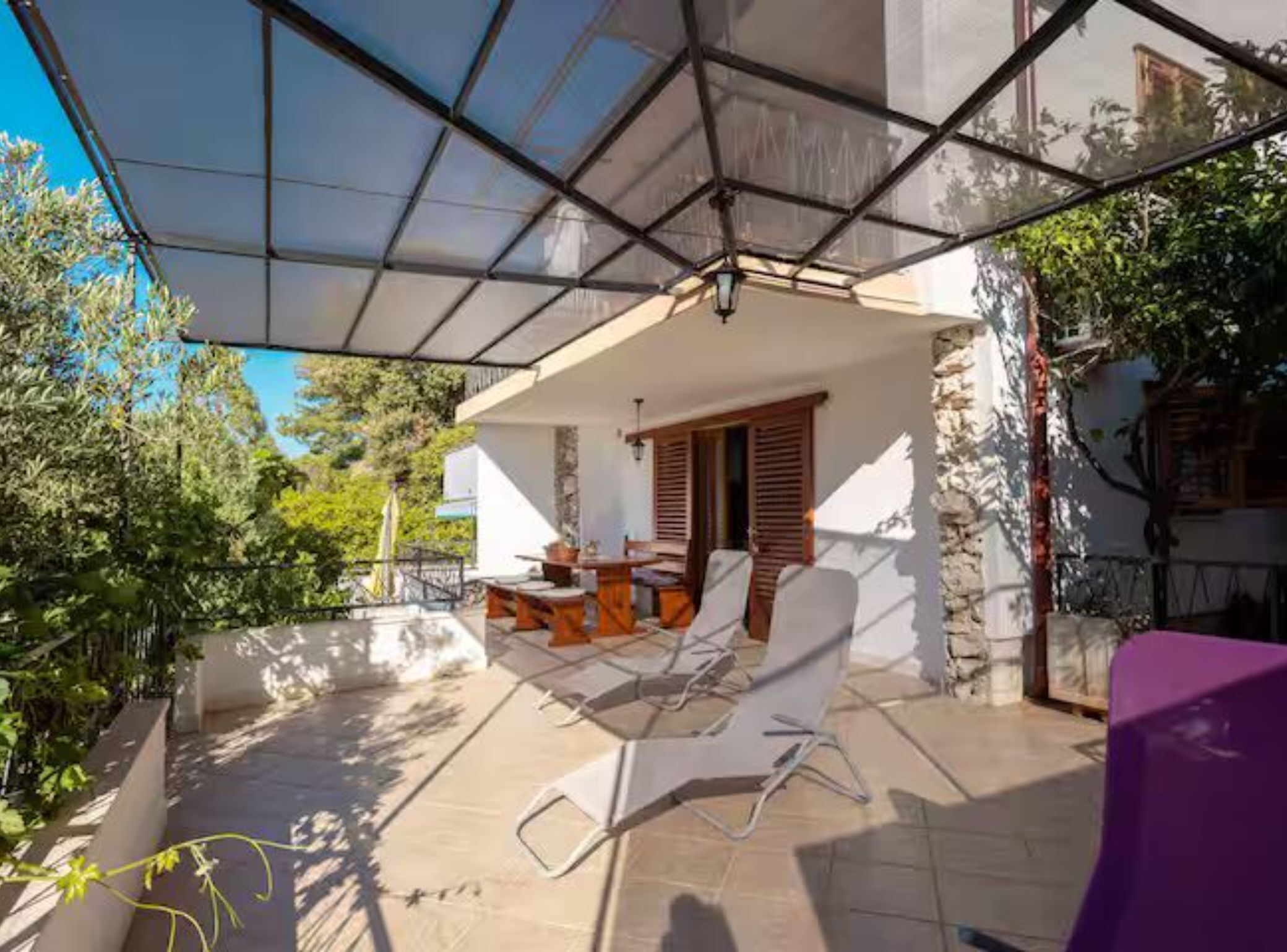 A covered outdoor patio with a table and chairs, a stone wall, and a view of greenery.
