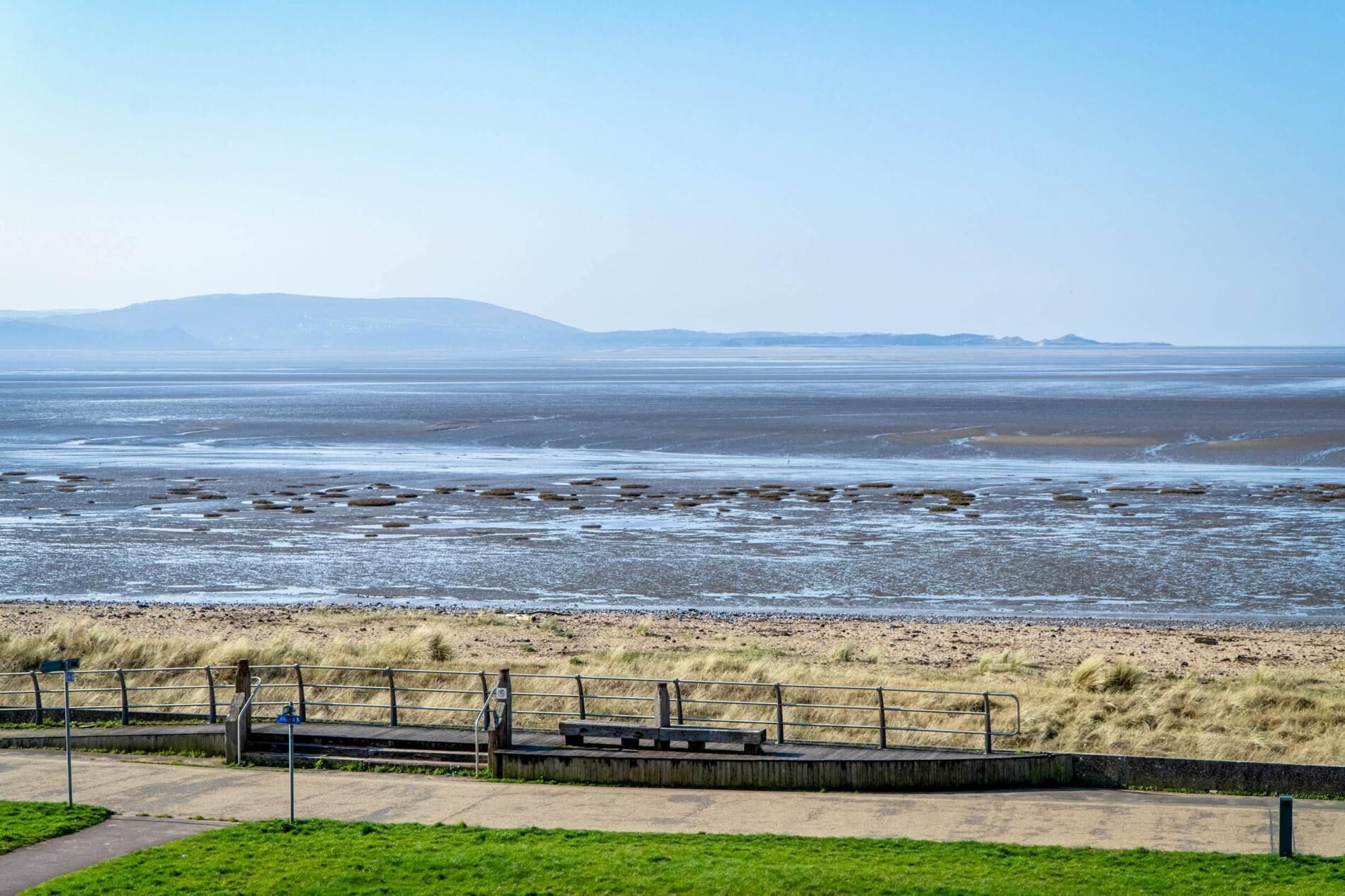 A coastal landscape with a sandy beach, a wooden boardwalk, and a grassy area.
