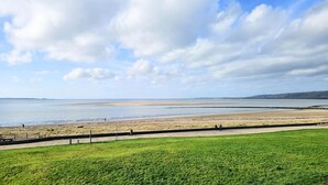 Un paisaje costero con un primer plano de hierba, una playa arenosa y un mar tranquilo bajo un cielo parcialmente nublado.