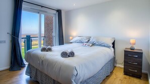 Un dormitorio con una cama grande, dos lámparas de noche, una mesita de madera y vistas a través de un ventanal corredero.