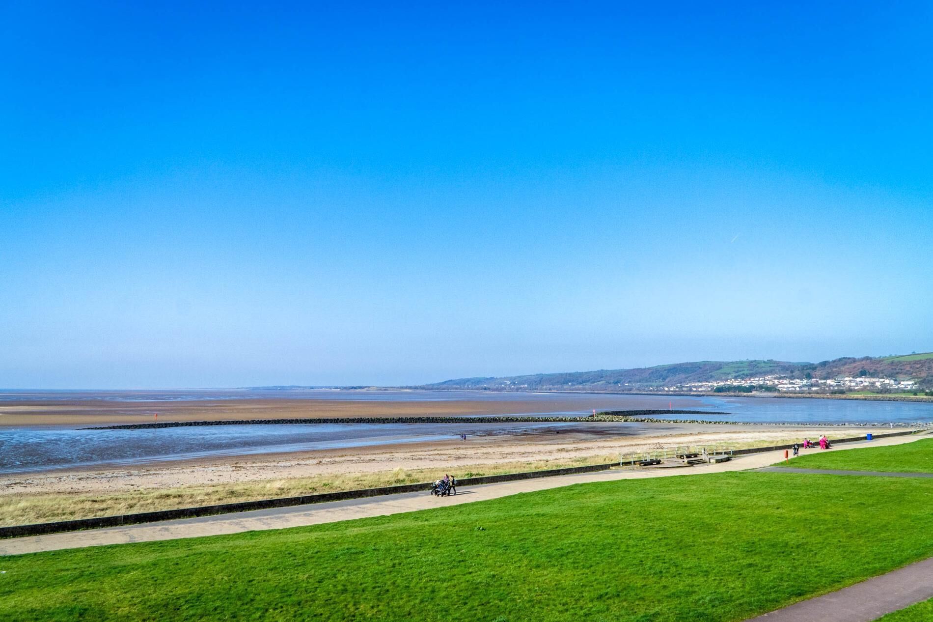 A coastal landscape with a clear sky, a sandy beach, a grassy area, and a distant town.