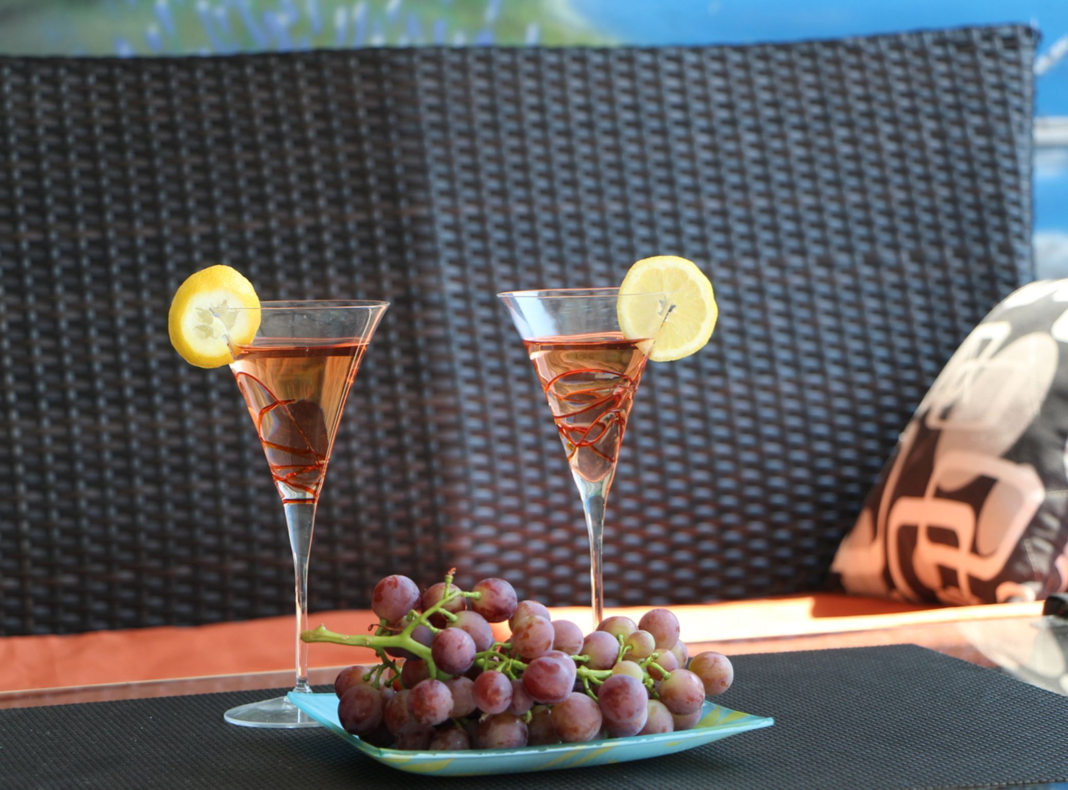 Duas taças de uma bebida rosa com rodelas de limão, um prato de uvas e um fundo de vime.
