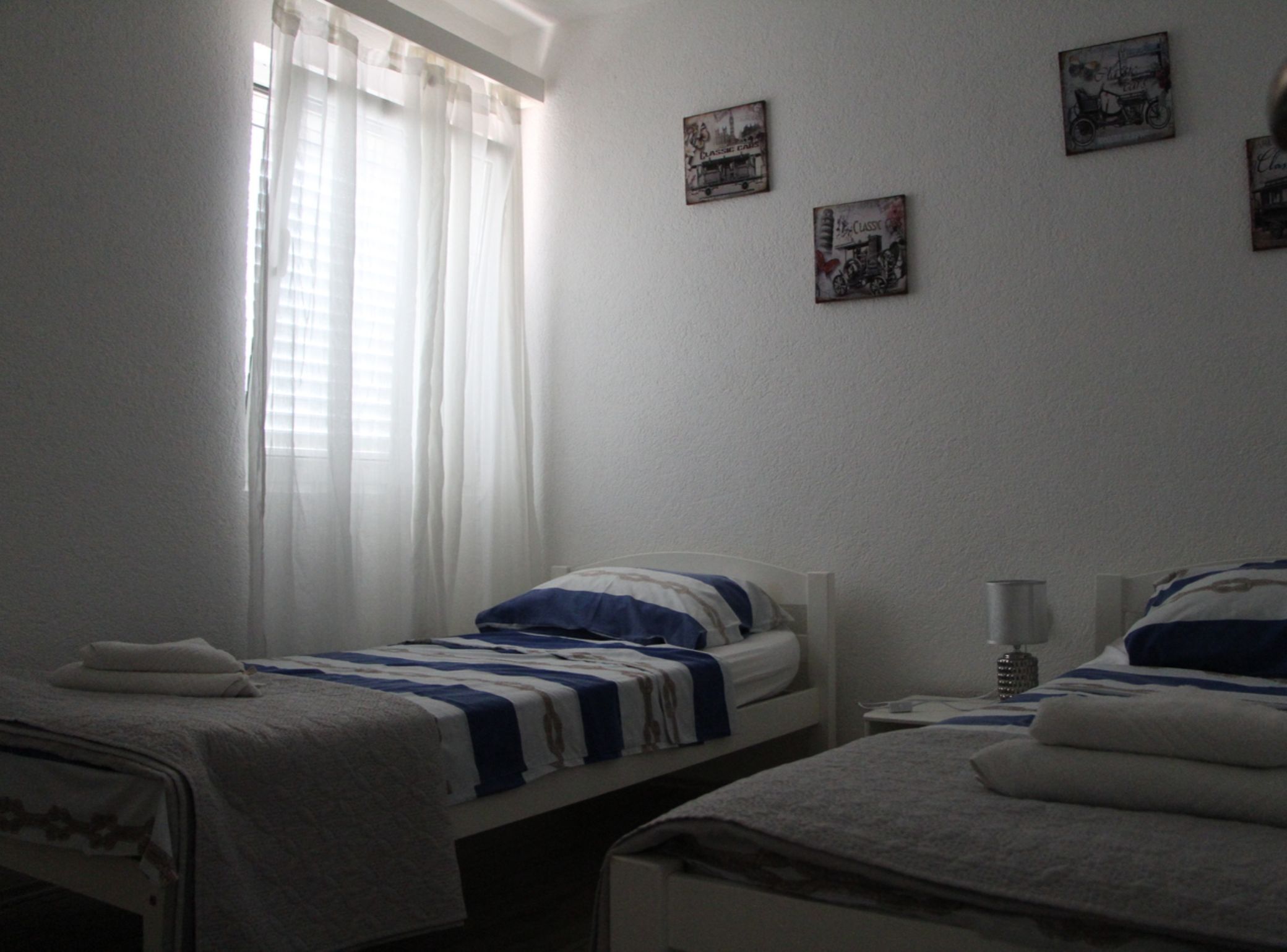 Ein Schlafzimmer mit zwei Einzelbetten, einem Nachttisch mit Lampe und einem Fenster mit durchsichtigen Vorhängen.