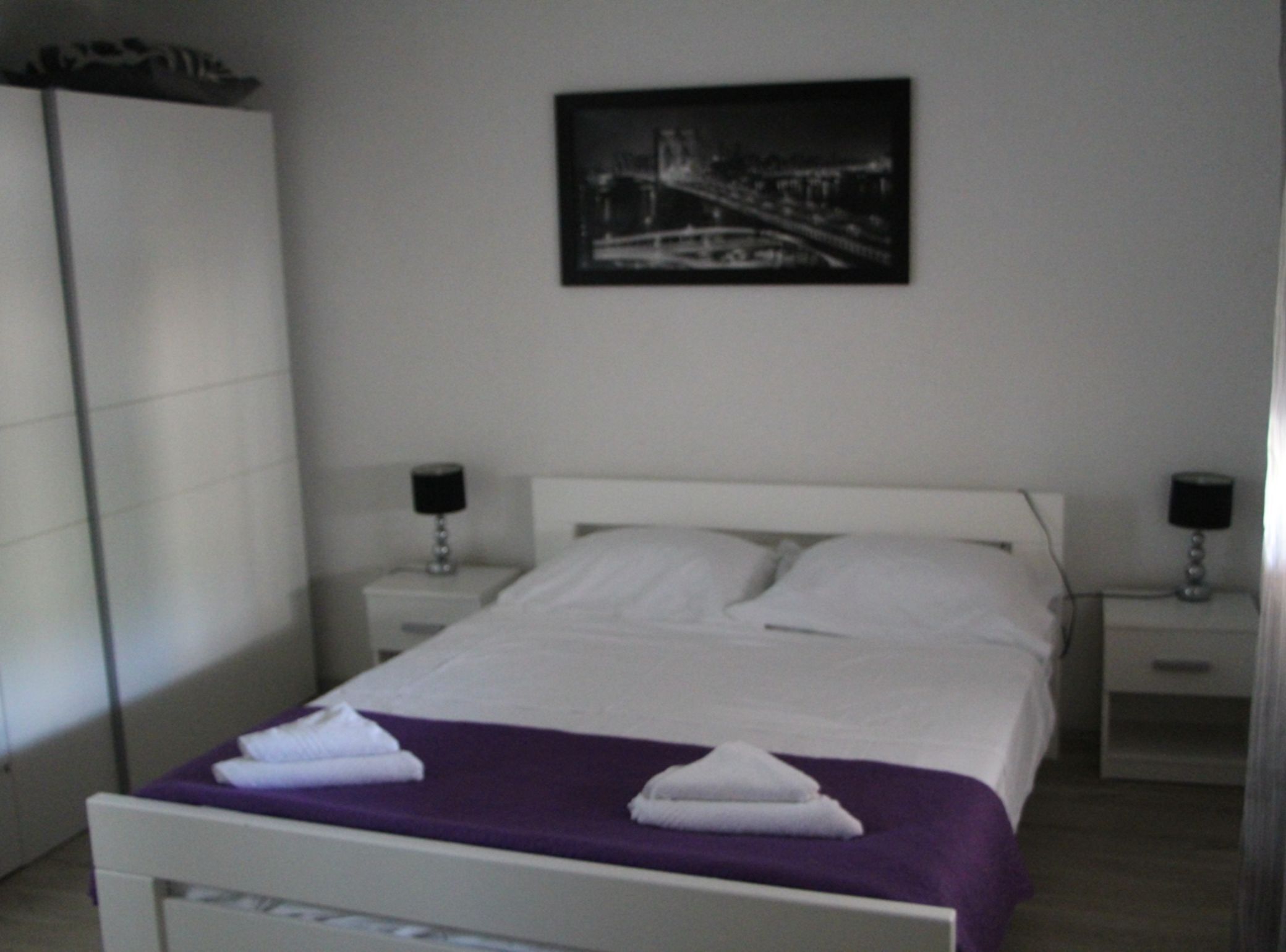 Una cama bien hecha con sábanas blancas y una colcha morada, dos mesitas de noche con lámparas, un armario blanco y un cuadro en la pared.