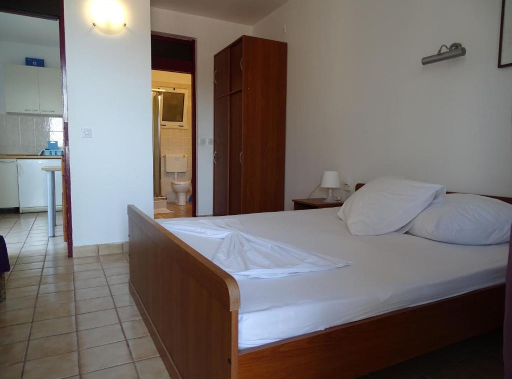 A bedroom with a wooden bed, a nightstand with a lamp, a wardrobe, and a view of a bathroom and kitchen.