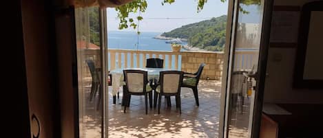 A room with a sliding glass door leading to an outdoor patio with a table and chairs, a pergola with hanging plants, and a view of the sea and hills.