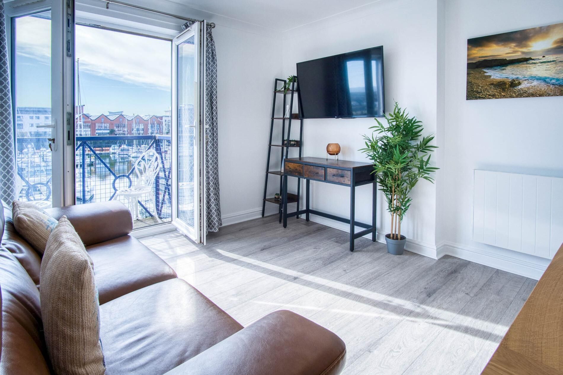 A modern living room with a sofa, a desk with a TV, a plant, and a balcony with a view.