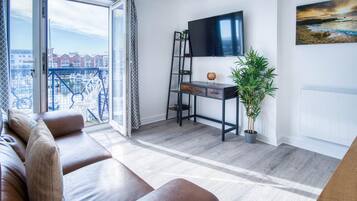A modern living room with a sofa, a desk with a TV, a plant, and a balcony with a view.