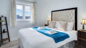 A neatly made bed with a blue throw blanket, two bedside lamps, and a city view through the window. - The Crows Nest - 1 Bed Apartment - Swansea Marina (Swansea)