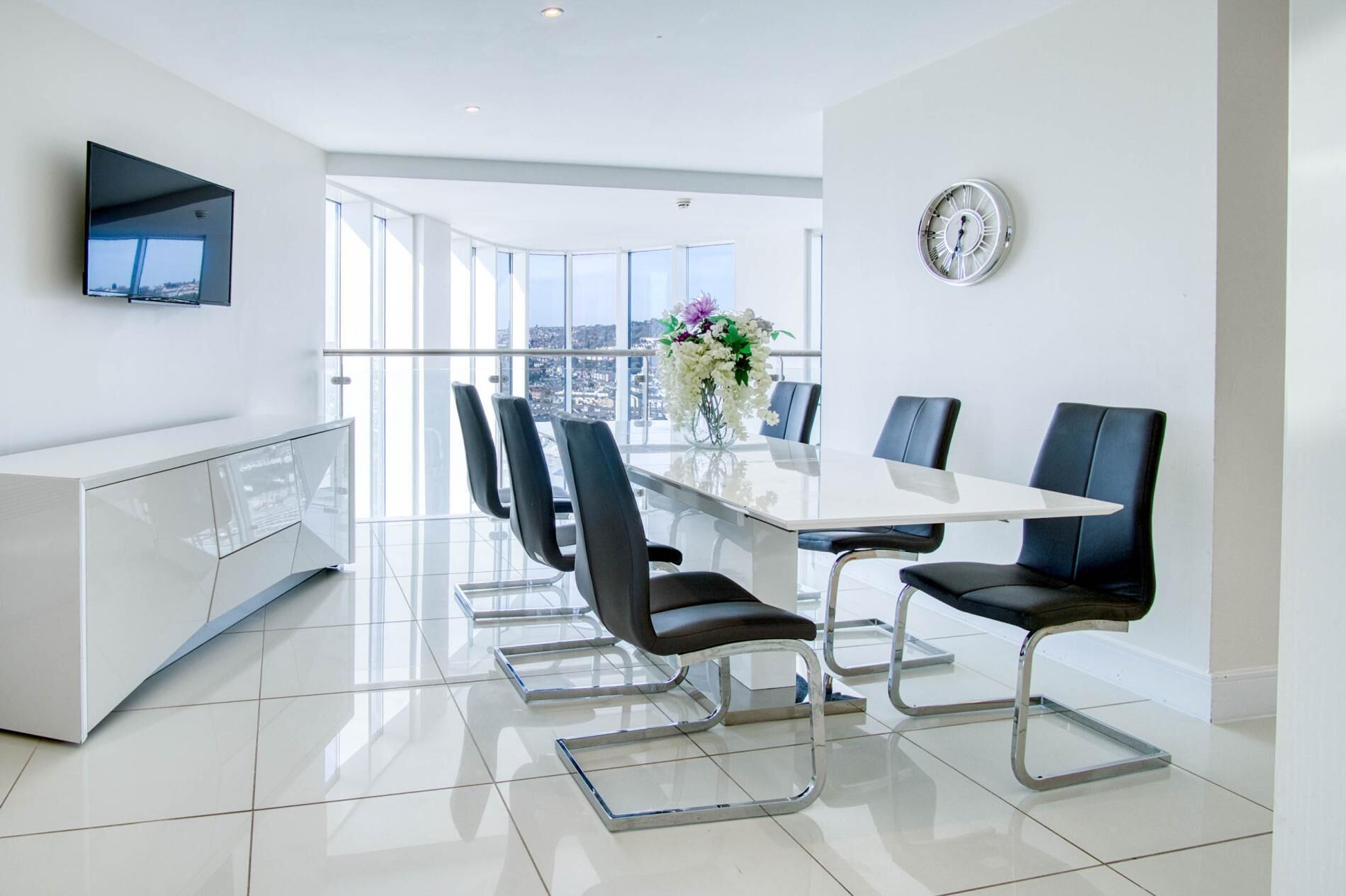 A modern meeting room with a glass table, black chairs, a white cabinet, a mounted TV, and a clock on the wall.