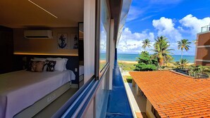 A balcony with a view of the beach and ocean, a bed with pillows, and a small table. - Studio Vista Mar Lateral 3 - By Almare (João Pessoa)