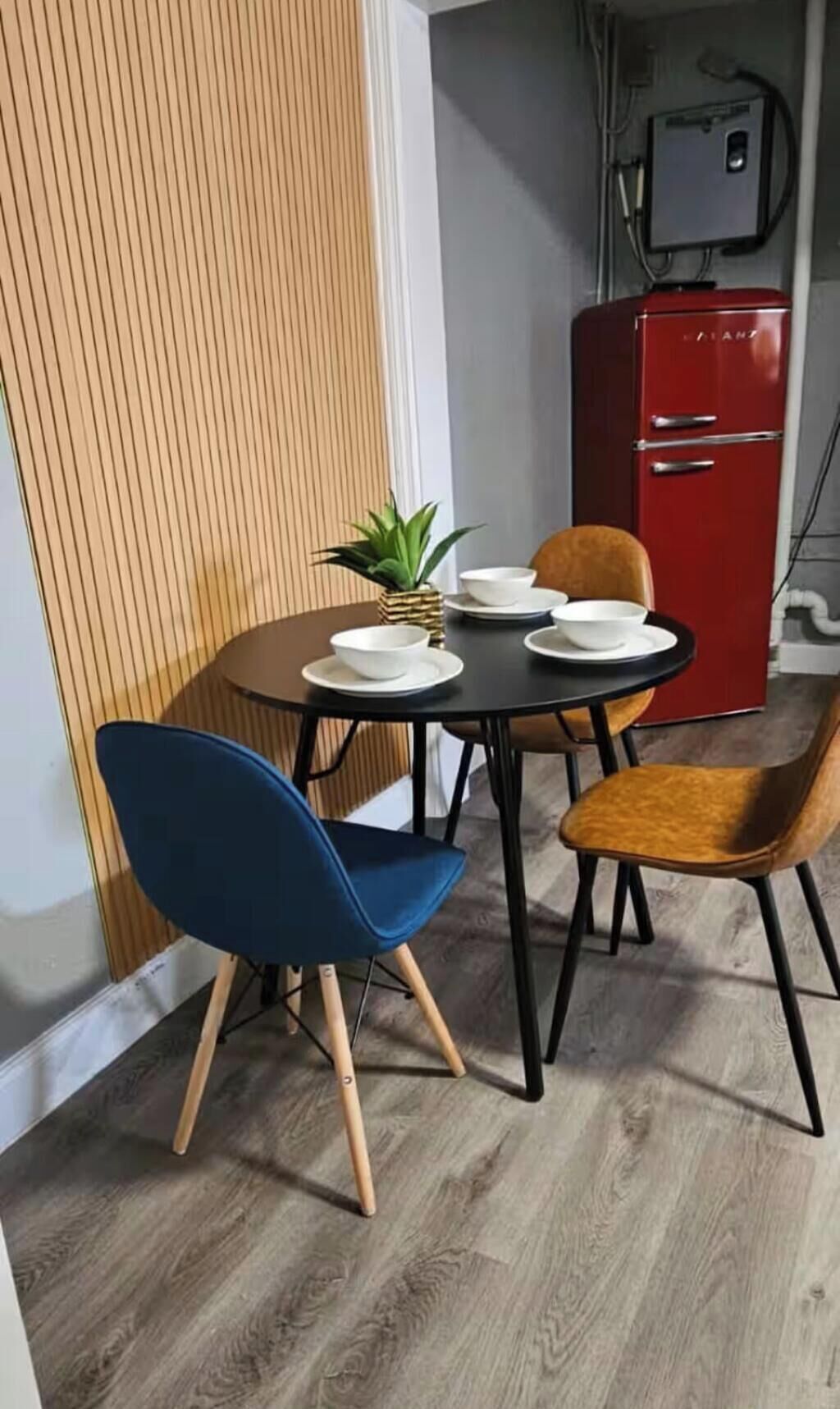 Un coin repas avec une table dressée pour quatre personnes, deux chaises bleues, deux chaises orange et un buffet rouge en arrière-plan.