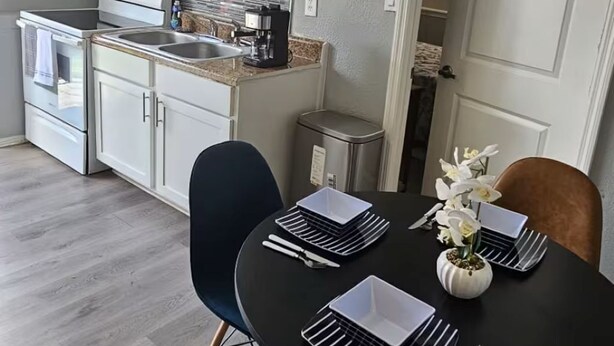 Ett kompakt kök med vita skåp, en mikrovågsugn och en spis. Här finns också en matplats med ett svart bord, vita tallrikar och en vas med blommor.
