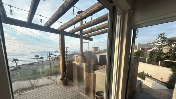 En balkong med trägolv, en pergola med hängande lampor, en krukväxt och utsikt över havet och byggnaderna.